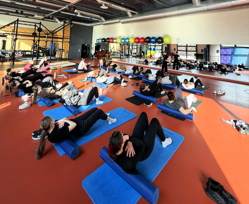 Odontologinės priežiūros studijų programos studentai sporto salėje atlieka tempimo ir stiprinimo pratimus, skirtus gerinti ergonomiką odontologijoje ir mažinti darbo metu patiriamą fizinę įtampą.