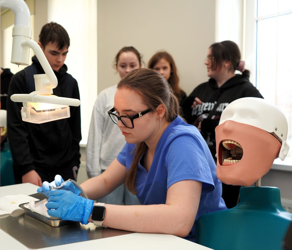 Burnos sveikatos edukacijos metu Kauno kolegijos Medicinos fakultete moksleiviai stebi odontologinės priežiūros studentės praktinį darbą su dantų modeliu ir odontologinėmis medžiagomis.