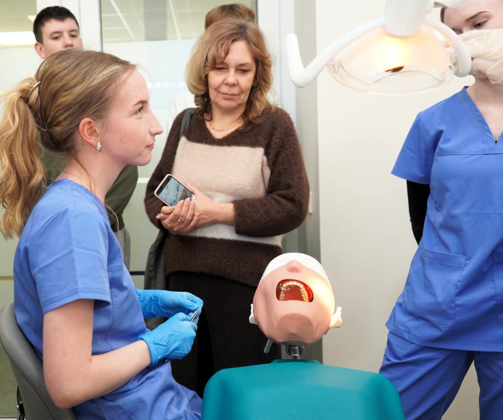 Burnos sveikatos edukacija Kauno kolegijos Medicinos fakultete – studentė demonstruoja odontologinių procedūrų įrankius ir darbą su dantų modeliu, o gimnazistai stebi praktinį užsiėmimą.
