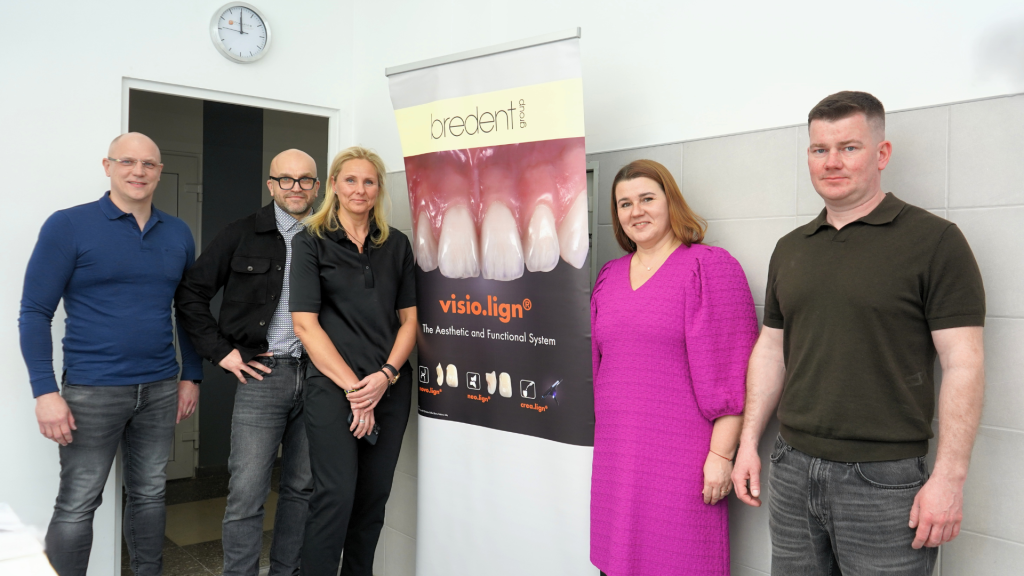 Dantų technologų profesinio meistriškumo konkurso vertinimo komisijos nariai, katedros vedėja ir socialiniai partneriai pozavo šalia renginio rėmėjo „Bredent group“ stendo.