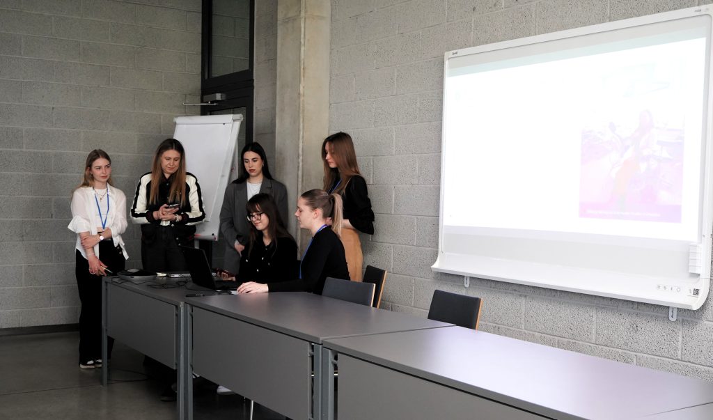 Studentės auditorijoje pristato grupinį darbą programos baigiamųjų pristatymų metu.