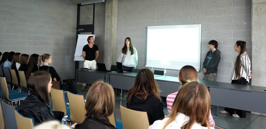 Programos dalyviai auditorijoje klausosi studentų pristatymo, rodomo interaktyvioje lentoje.