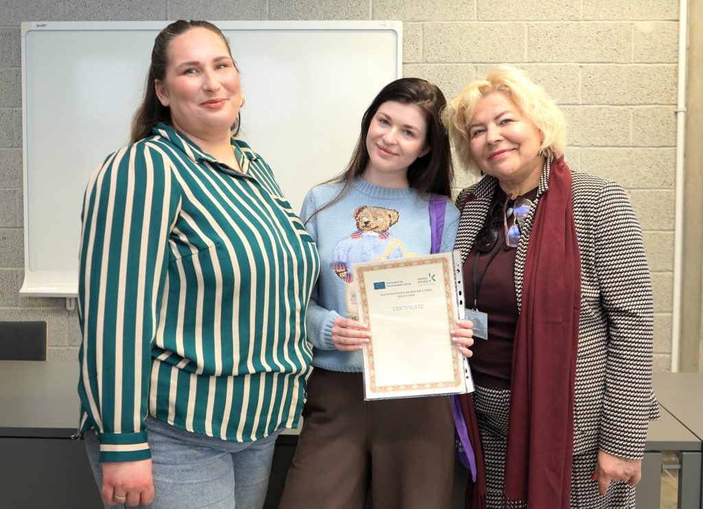 BIP dalyvė su sertifikatu fotografuojasi kartu su dėstytojomis po programos veiklų.