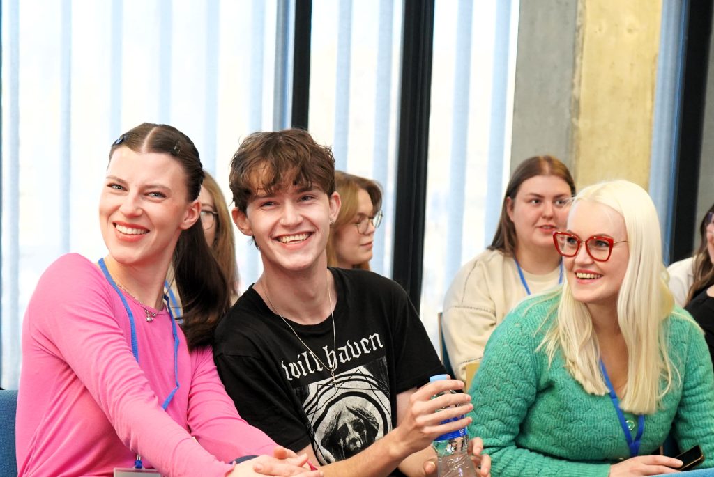 Programos studentai sėdi auditorijoje, bendrauja ir šypsosi programos užsiėmimų metu.