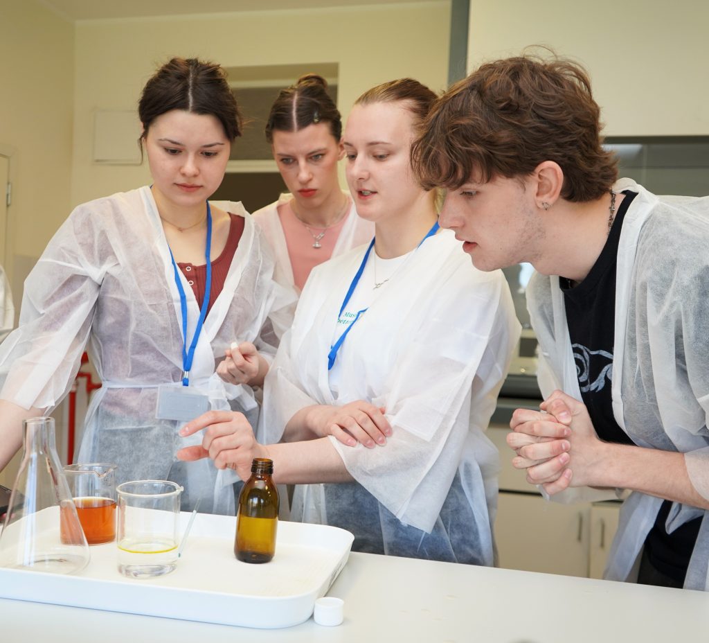 „Wellbeing from Nature“ dalyviai laboratorijoje stebi praktinį procesą prie stalo su stikliniais indais ir priemonėmis.