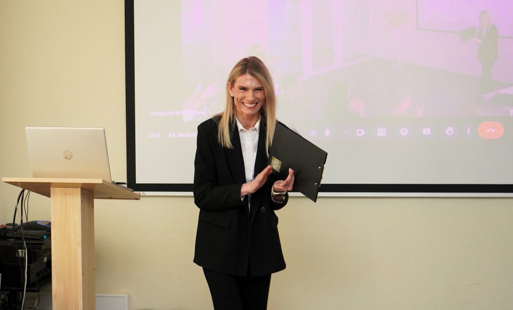 Konferencijos „Mokslas ir studijos: 2026“ Medicinos fakulteto dekanė scenoje šypsodamasi laiko aplanką, šalia matomas projektorius ir kompiuteris.