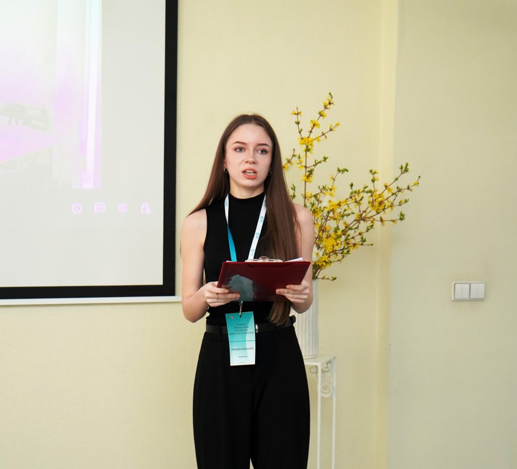 Studentė skaito pranešimą konferencijoje, rankose laikydama užrašus ir stovėdama prie ekrano.