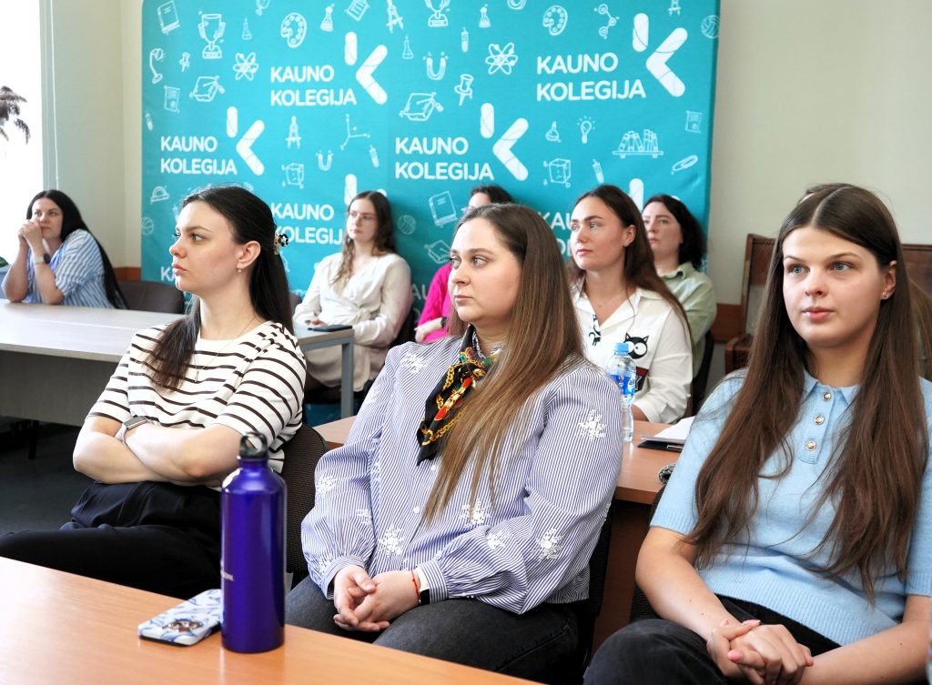 Auditorijoje sėdintys studentai klausosi pranešimų konferencijoje, fone matomas Kauno kolegijos stendas.