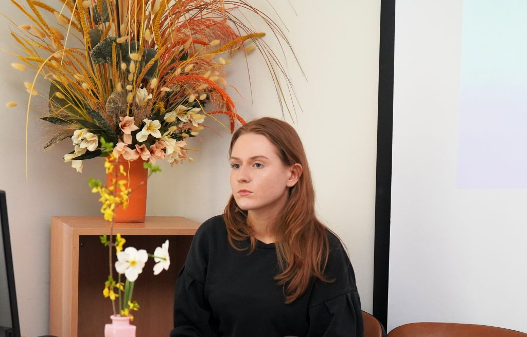 Studentė ruošiasi skaityti pranešimą konferencijoje „Mokslas ir studijos: 2026“, sėdėdama šalia dekoratyvinių gėlių kompozicijos.