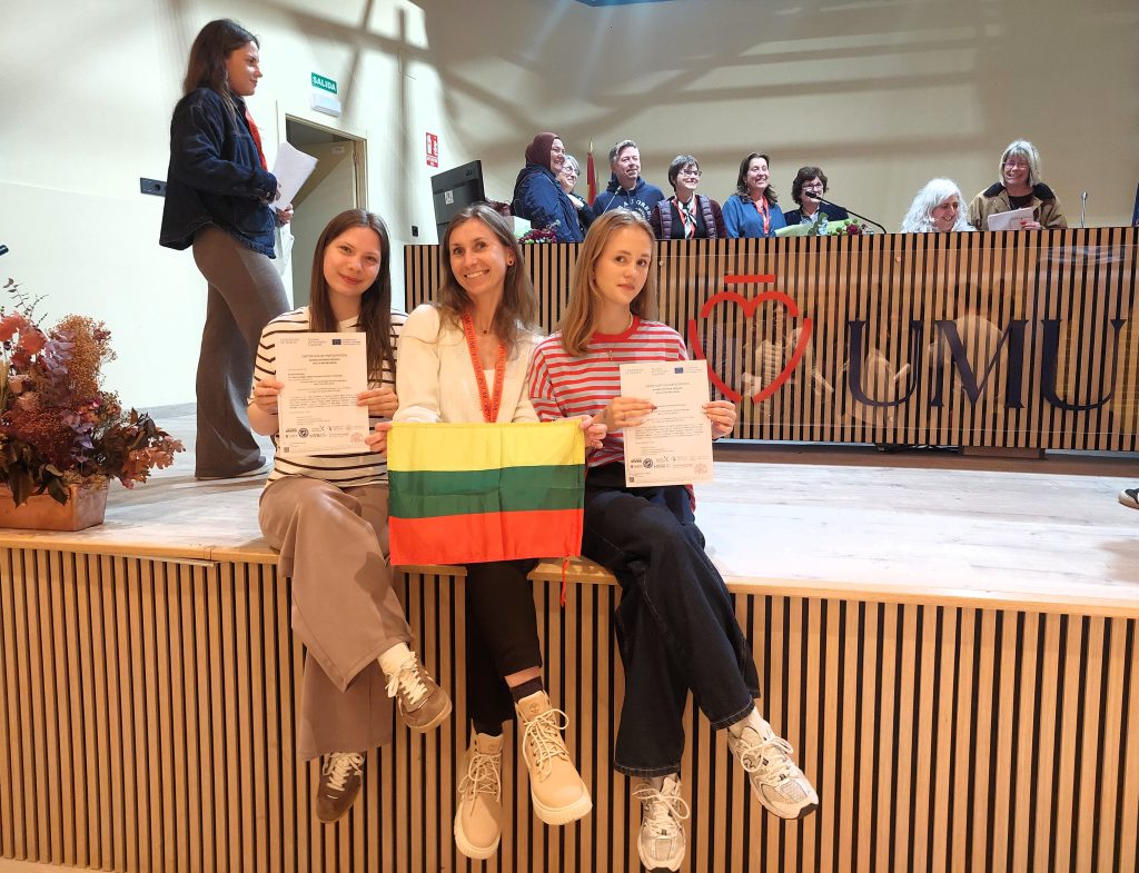 Tarptautinio renginio dalyvės studentės kartu su reabilitacijos katedros dėstytoja pozuoja nuotraikai su pažymėjimais ir Lietuvos vėliava.