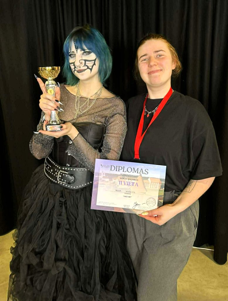 Kosmetologijos studenčių pasirodymas Lietuvos kirpėjų ir grožio specialistų atvirame čempionate „Kaunas Beauty 2026“. Nuotraukoje dvi merginos pozuoja prie juodos užuolaidos, viena jų laiko taurę, o kita rankose laiko diplomą. Modelis išsiskiria ryškiu meniniu makiažu ir tamsiu sceniniu įvaizdžiu.