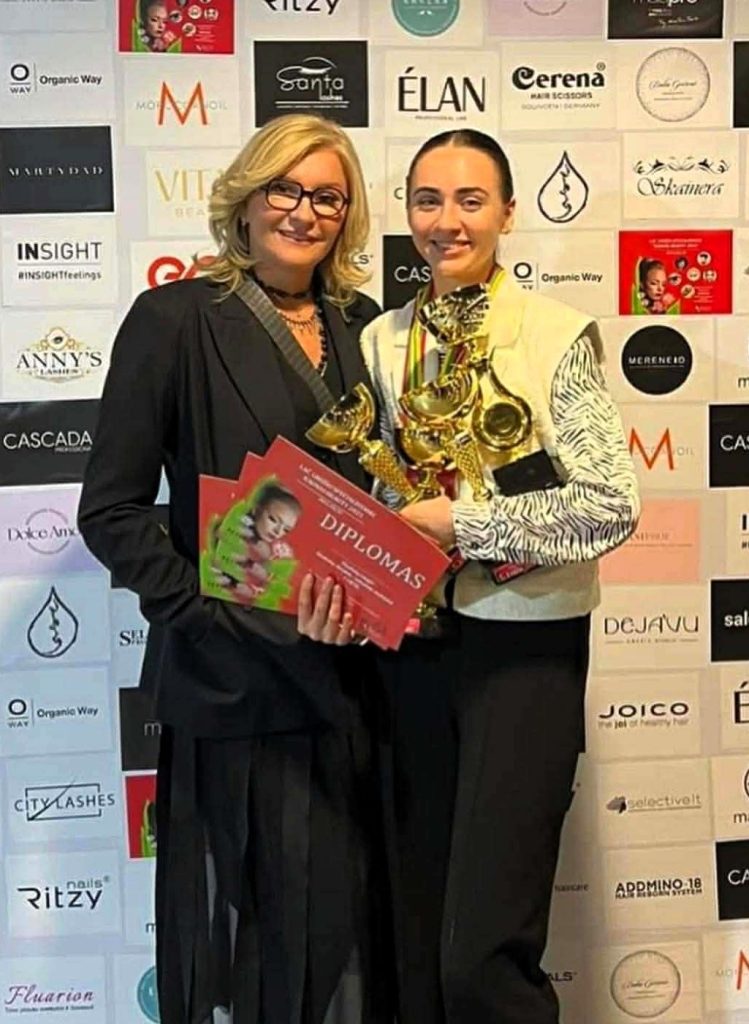 Nuotraukoje dvi moterys stovi prie rėmėjų sienelės, viena jų rankose laiko kelias taures ir diplomą. Abi pozuoja po apdovanojimų ceremonijos.