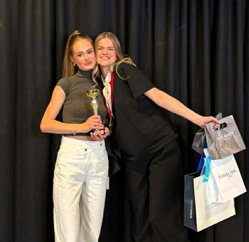 Kosmetologijos studenčių pasirodymas Lietuvos kirpėjų ir grožio specialistų atvirame čempionate „Kaunas Beauty 2026“. Nuotraukoje dvi merginos pozuoja apsikabinusios prie juodos užuolaidos. Viena jų rankose laiko taurę, kita rankoje turi kelis dovanų maišelius.