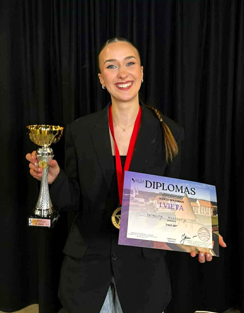 Nuotraukoje mergina prie juodos užuolaidos pozuoja su taure, diplomu ir medaliu ant kaklo. Ji šypsosi ir laiko apdovanojimus rankose.