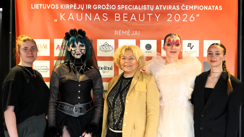 Kosmetologijos studenčių pasirodymas Lietuvos kirpėjų ir grožio specialistų atvirame čempionate „Kaunas Beauty 2026“, kur prie renginio sienelės pozuoja studentės, dėstytoja ir du modeliai su sceniniu makiažu.