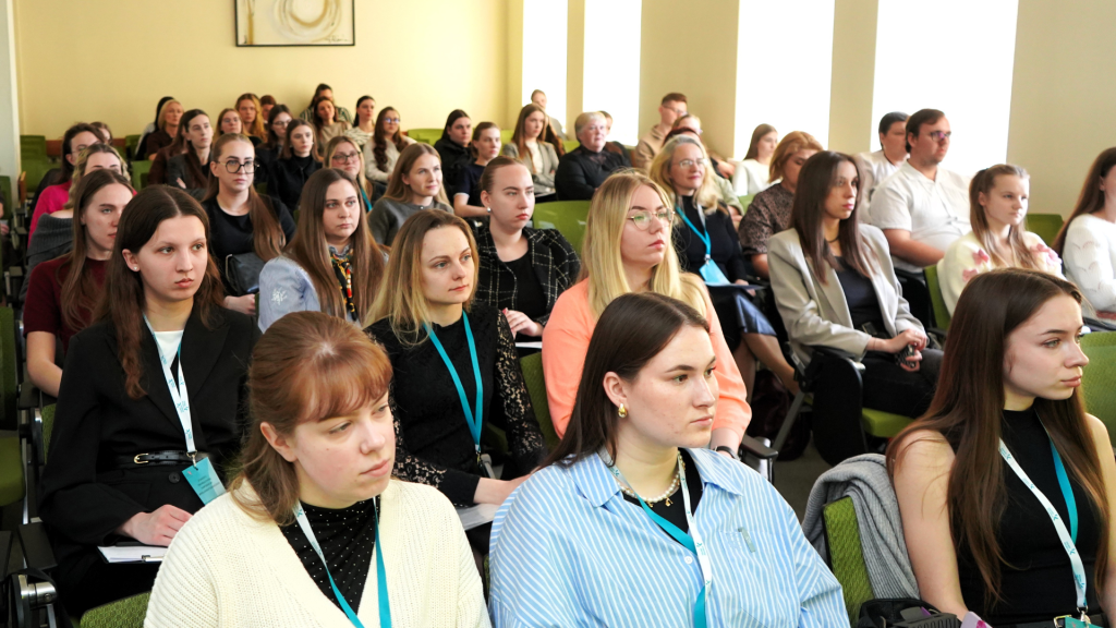 Studentų auditorija konferencijoje „Mokslas ir studijos: 2026“, kurioje gausus būrys klausytojų atidžiai klausosi pranešimų, sėdi auditorijoje su užrašų knygomis ir konferencijos kortelėmis.