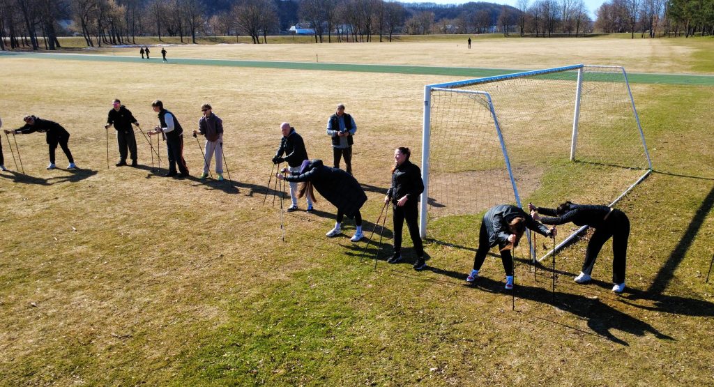 Šiaurietiškojo ėjimo paskaitos dalyviai atlieka tempimo pratimus stadione su ėjimo lazdomis