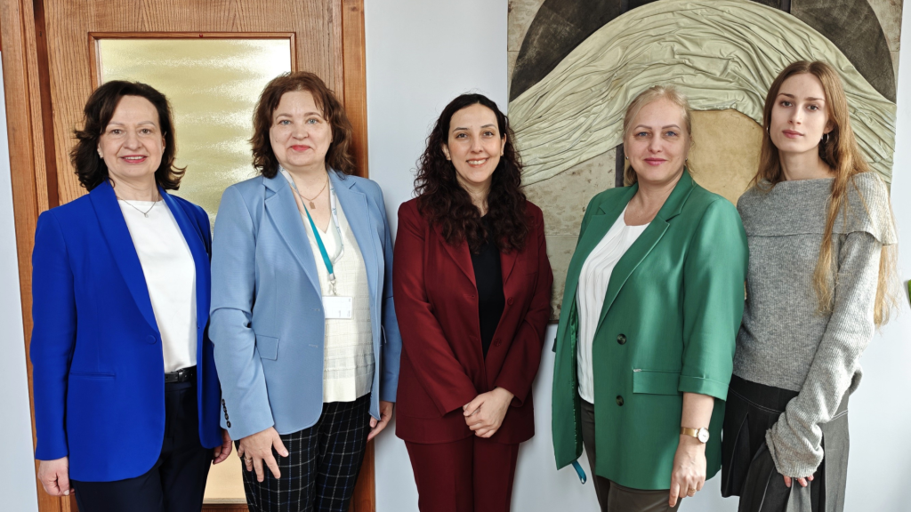 Pediatrinės slaugos dėstytoja iš Turkijos su Medicinos fakulteto Slaugos katedros atstovėmis stovi patalpoje ir pozuoja bendrai nuotraukai.