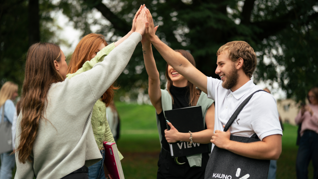 Kauno kolegijos studentai lauke džiaugsmingai sveikinasi pakeltomis rankomis, simbolizuodami bendrystę, studijų patirtį ir pasirengimą darbo rinkos poreikiams.