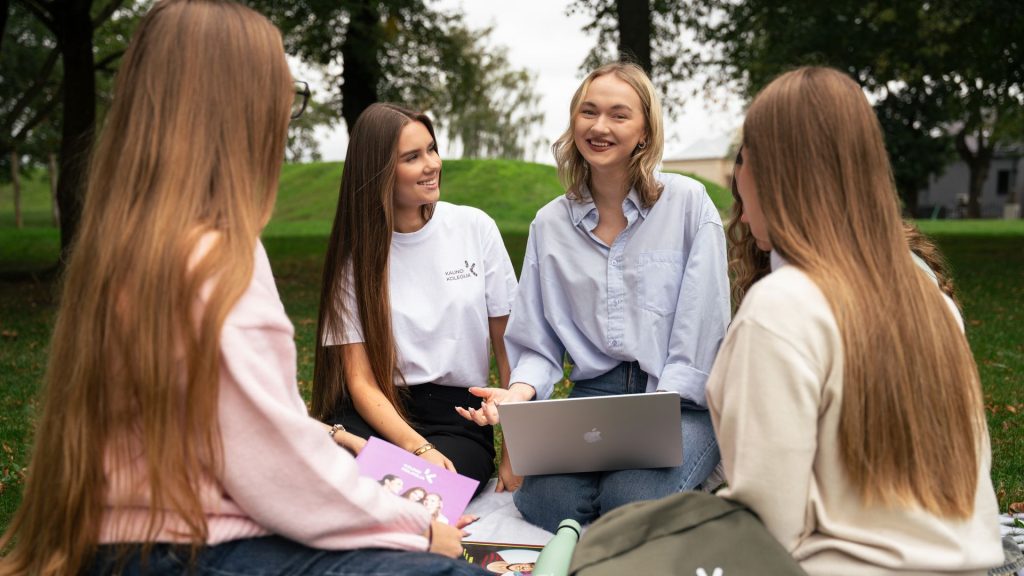 Keturi Kauno kolegijos studentai diskutuoja lauke, ant pledo, su nešiojamuoju kompiuteriu.