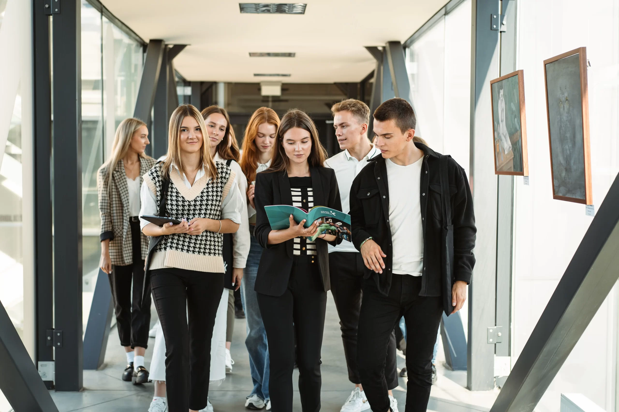 Studentų grupė eina stikliniu koridoriumi, nešini planšete ir aptarinėja informacinį lankstinuką.