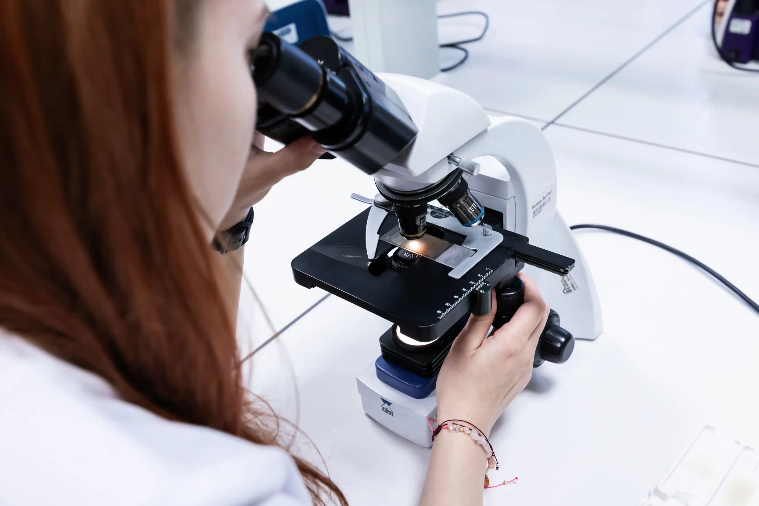 
Studentė su laboratoriniu chalatu atidžiai reguliuoja mikroskopą tirdama mėginį.