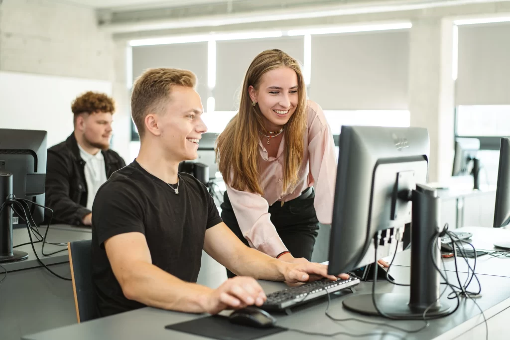 Besišypsantys studentai bendradarbiauja prie kompiuterio modernioje auditorijoje.