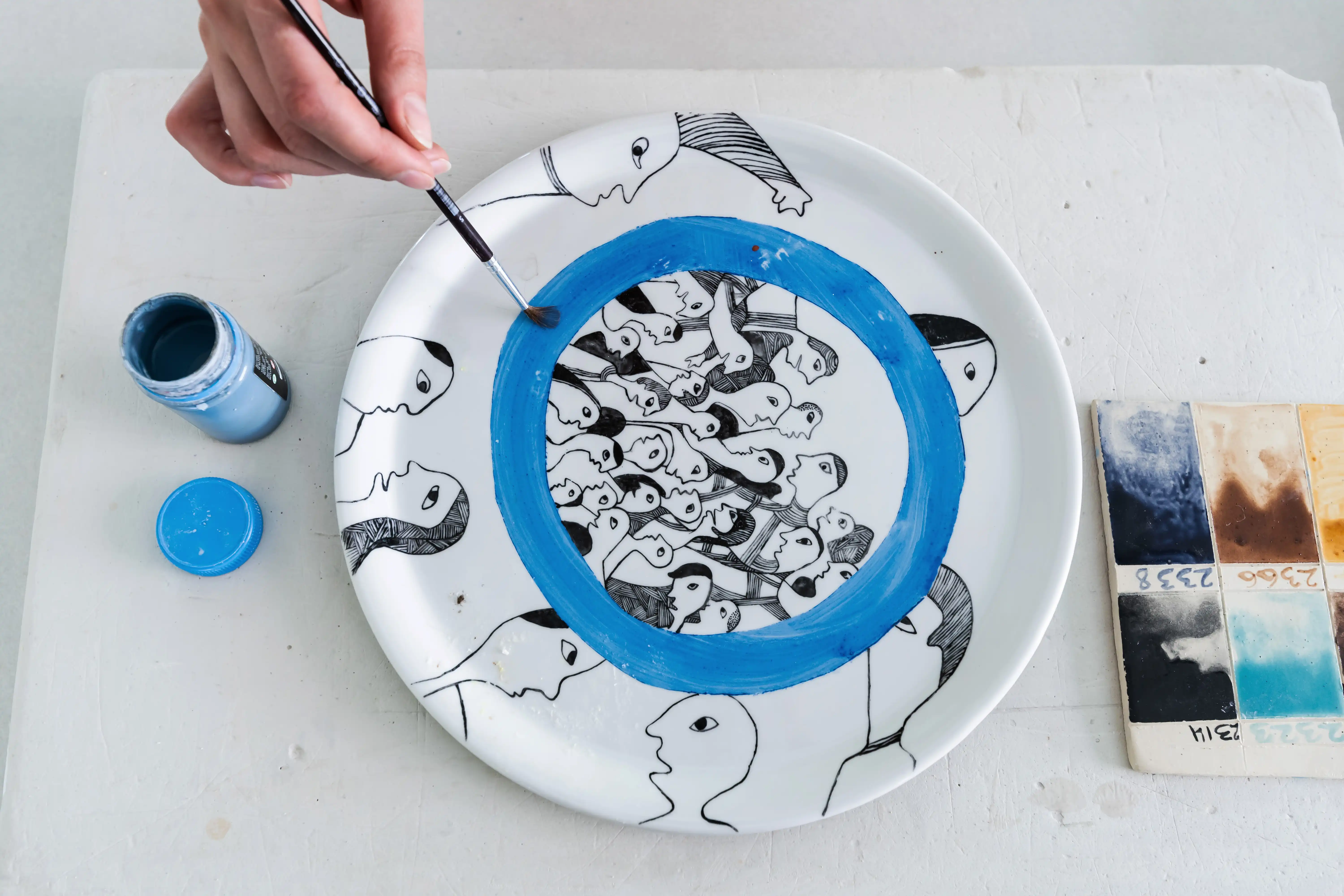 Studentas teptuku tapo mėlyną ratą ant keraminės lėkštės, dekoruotos stilizuotais veidų piešiniais.