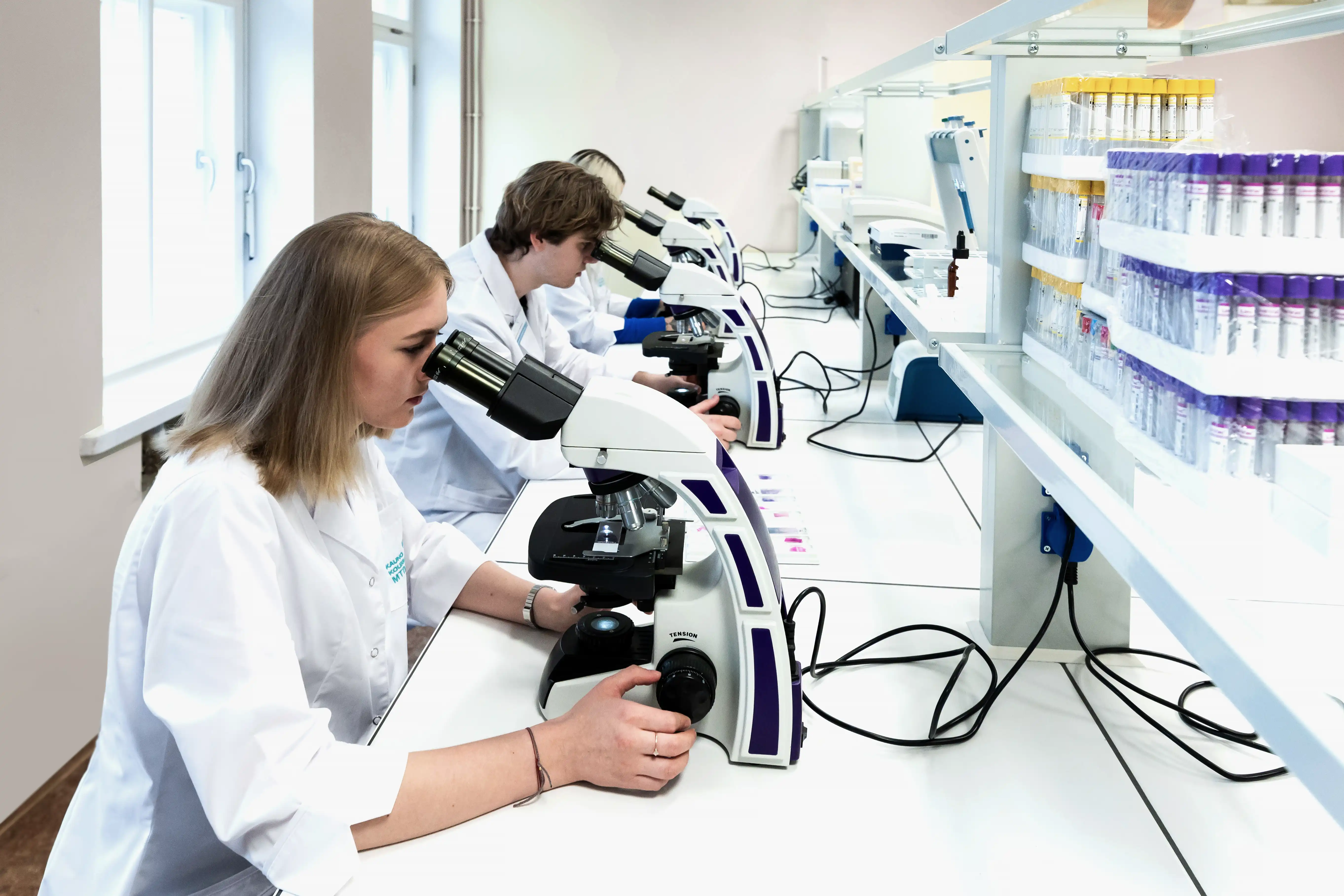 Studentai su baltais chalatais modernioje laboratorijoje tiria mėginius mikroskopais.