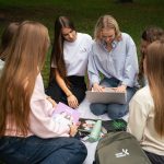 Šešios merginos sėdi ratu ant pledo parke ir kartu žiūri į nešiojamą kompiuterį; šalia padėtos knygos ir kuprinė.