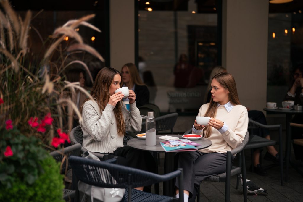 Dvi jaunos moterys sėdi lauko kavinėje prie nedidelio staliuko, geria kavą iš baltų puodelių ir kalbasi; ant stalo matyti žurnalas, vandens butelis ir lėkštelės, o aplink – kiti lankytojai bei augalai.