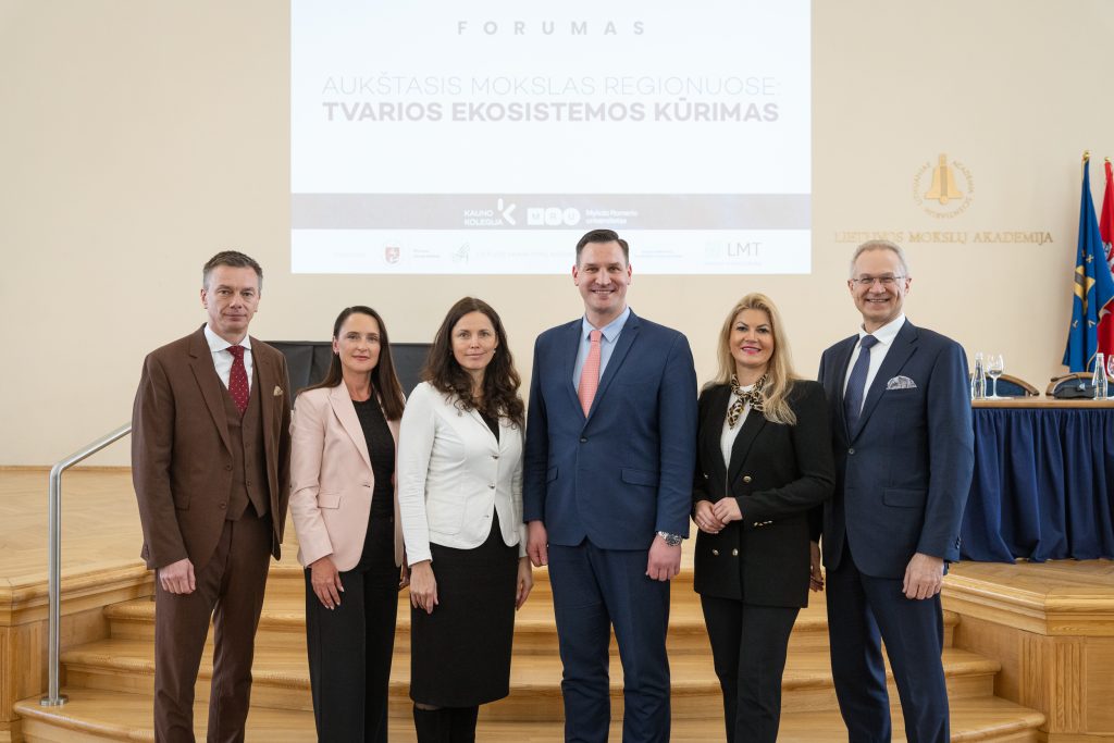 Šeši oficialiai apsirengę asmenys stovi scenoje per forumą apie aukštąjį mokslą regionuose, už jų matomas renginio pristatymo ekranas.