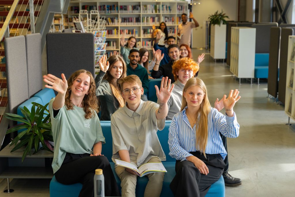 Studentų grupė modernioje bibliotekoje sėdi ant sofų ir draugiškai mojuoja, laikydami knygas ir bendraudami tarpusavyje.