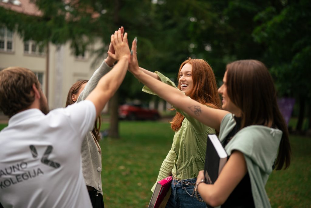 Grupė jaunų žmonių lauke, žalioje aplinkoje, džiaugsmingai susiduria rankomis „penketuku“, šypsosi ir laiko knygas, tarsi po sėkmingos veiklos ar mokymosi akimirkos.