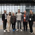 Šeši žmonės stovi greta modernaus pastato vestibiulyje su stiklinėmis sienomis ir metalinėmis konstrukcijomis; visi vilki kasdienius-verslo drabužius ir žiūri į kamerą.