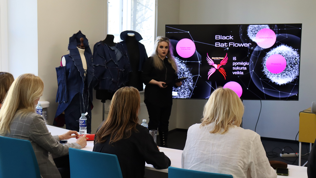 Moteris stovi prie ekrano ir pristato projektą „Black Bat Flower“, o priešais ją sėdintys klausytojai stebi prezentaciją; šalia matomi tamsūs drabužių dizaino modeliai ant manekenų.