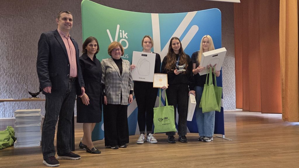 Grupė žmonių pozuoja po apdovanojimų ceremonijos. Trys studentės laiko diplomą bei prizus prie VIKO renginio sienelės.