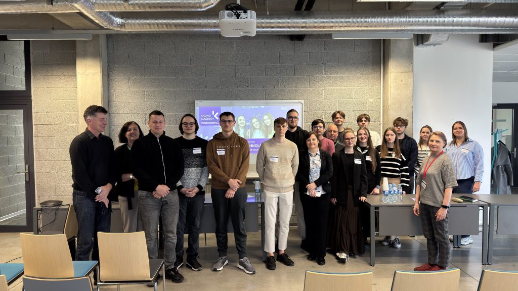 Grupinė nuotrauka modernioje auditorijoje – apie 18 žmonių stovi priekyje priešais projektoriaus ekraną, ant kurio rodoma prezentacija. Dalyviai vilki kasdieniais drabužiais, kai kurie turi vardines korteles. Patalpoje matomos betoninės sienos, atviros ventiliacijos sistemos lubose, stalai su vandens buteliukais ir kėdės pirmame plane.