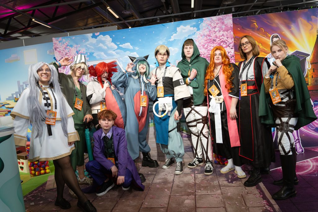 Gausi Kauno kolegijos studentų ir atstovų grupė pozuoja populiariosios kultūros renginyje (pvz., „Comic Con Baltics“). Dalis dalyvių vilki detalius „cosplay“ kostiumus, įkūnydami anime ir žaidimų personažus. Grupė stovi priešais dideles iliustruotas sieneles su sakurų ir fantastinių peizažų motyvais, visi ryši parodos dalyvių („Exhibitor“) korteles.