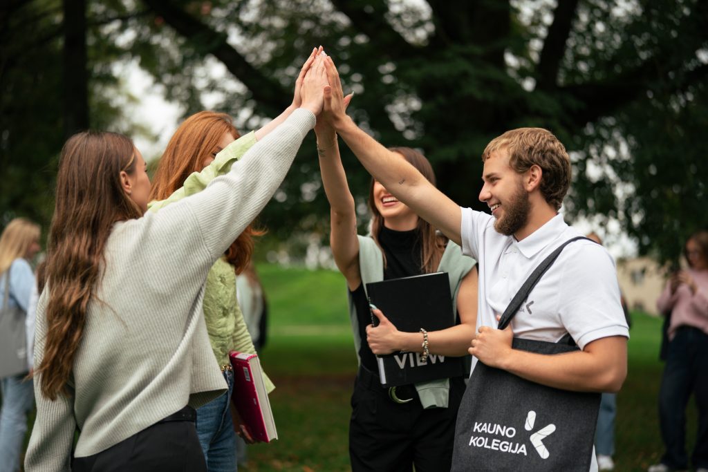Grupė jaunų žmonių lauke susidaužia delnais „high five“ gestu, šypsosi ir bendrauja žalio parko aplinkoje.