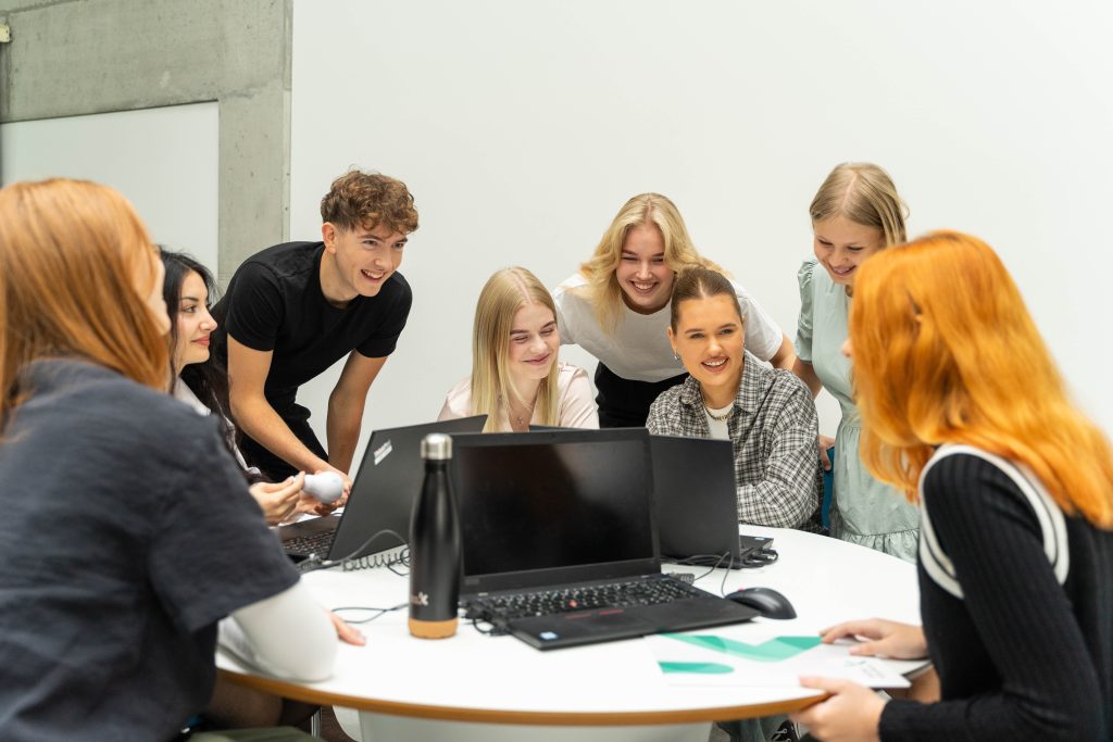 Besišypsantys Kauno kolegijos studentai bendradarbiauja ir diskutuoja prie nešiojamųjų kompiuterių šviesioje auditorijoje.