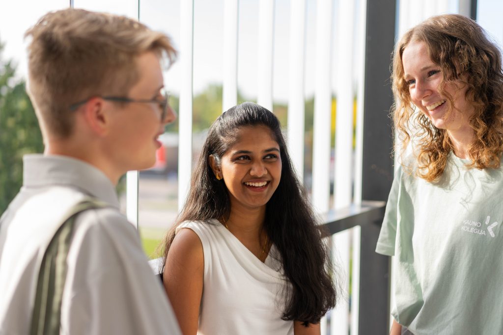 Trys studentai stovi universitete, šypsosi ir bendrauja tarpusavyje.