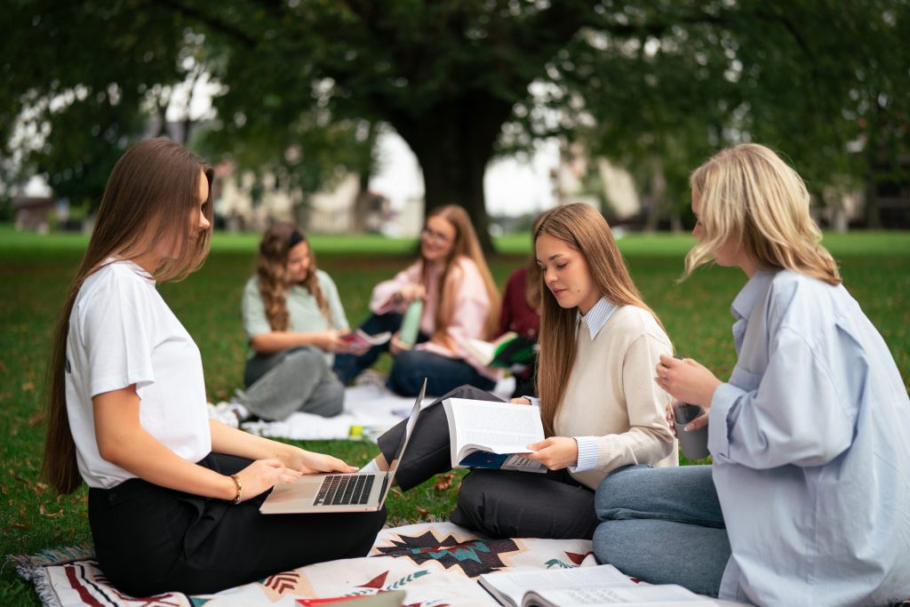 Kelios studentės sėdi ant pledo parke po medžiais, skaito knygas, dirba kompiuteriu ir bendrauja lauke.