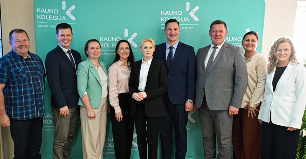 Devynių asmenų grupė (penkios moterys ir keturi vyrai), vilkintys dalykinę aprangą, pozuoja bendrai nuotraukai priešais turkio spalvos sienelę su Kauno kolegijos Alytaus fakulteto logotipais. Nuotraukos centre stovi Kauno kolegijos direktorius dr. Andrius Brusokas.