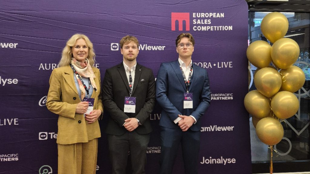Trys renginio dalyviai su oficialiomis akreditacijomis pozuoja prie violetinės reklaminės sienelės su užrašu „European Sales Competition“. Kairėje stovi moteris smėlio spalvos kostiumu ir skarele, centre – vyras juodu kostiumu, dešinėje – vyras mėlynu kostiumu. Dešinėje nuotraukos pusėje matyti aukso spalvos balionų kompozicija.