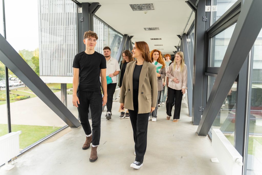 Būrys besišypsančių Kauno kolegijos studentų žengia moderniu stikliniu pereinamuoju tiltu, jungiančiu pastatus. Pirmame plane matomi besikalbantys jaunuolis ir mergina su švarku, o už jų seka kiti studentai, nešini studijų medžiaga.