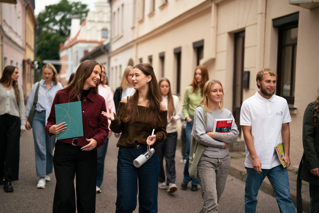 Grupė jaunų žmonių eina siaura senamiesčio gatve. Priekyje dvi merginos šypsodamosi kalbasi, viena rankoje neša aplanką, kita – gertuvę. Aplink jas eina kiti studentai su knygomis ir kuprinėmis, o fone matyti šviesių spalvų pastatai.