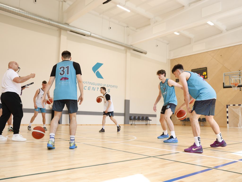 Kauno kolegijos krepšinio komandos nariai treniruojasi Sveikatingumo ir sporto centre. Nuotraukoje matomi turkio spalvos sportinę aprangą vilkintys studentai, varinėjantys krepšinio kamuolius, ir treneris baltais marškinėliais, stebintis procesą. Fone ant sienos matomas Kauno kolegijos logotipas ir užrašas „Sveikatingumo ir sporto centras“.