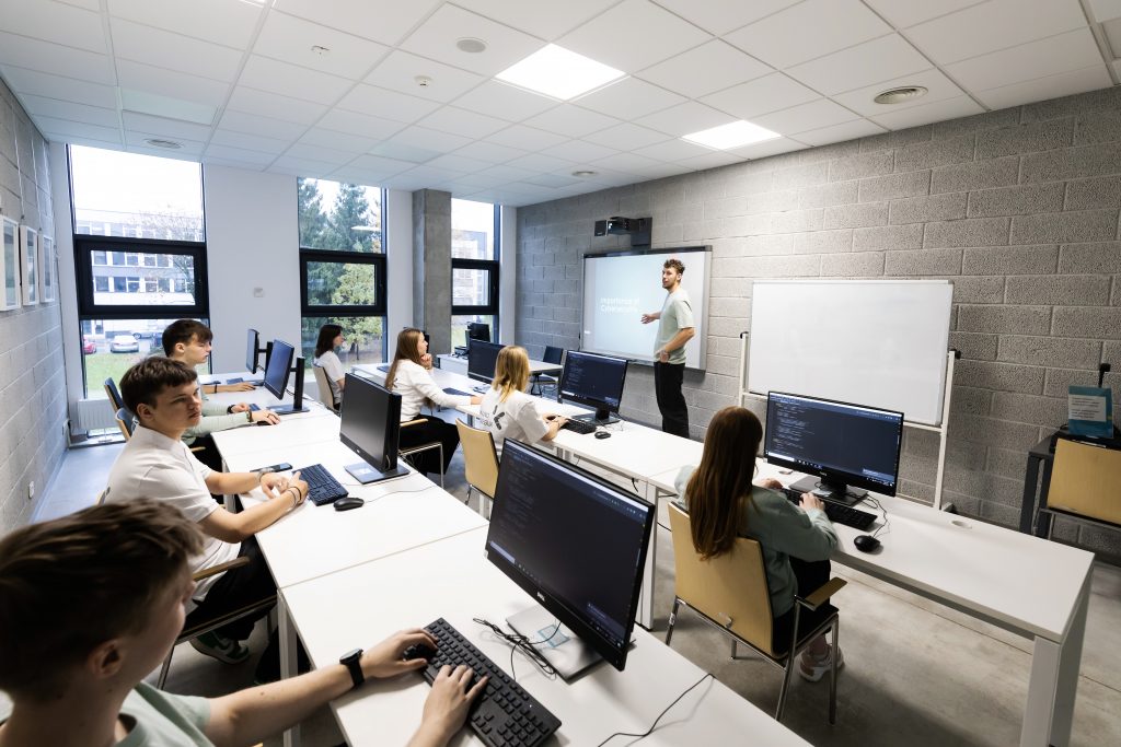 Programavimo klasėje dėstytojas stovi prie interaktyvios lentos ir veda pamoką, o keli mokiniai sėdi prie stalų su stacionariais kompiuteriais ir dirba su programavimo kodu ekranuose. Šviesi, moderni mokymo erdvė su dideliais langais ir balta lenta.