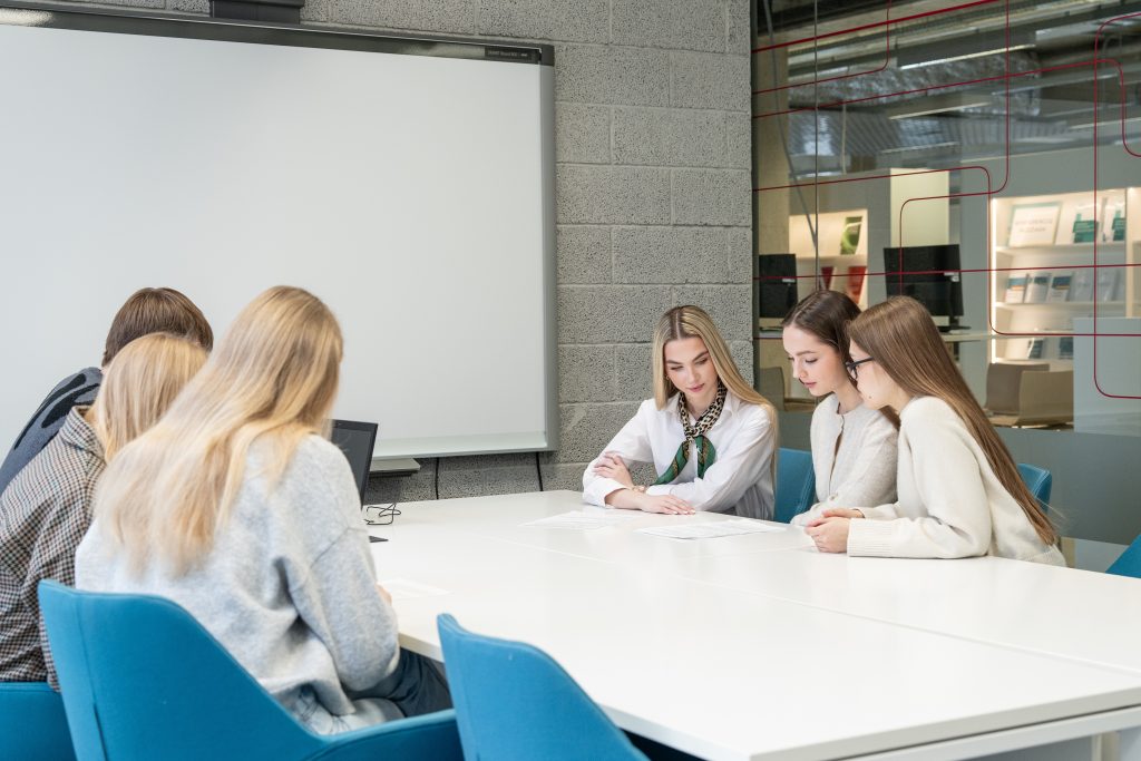 Šviesioje posėdžių ar mokymų salėje prie balto stalo sėdi grupė jaunų žmonių. Trys dalyvės dešinėje pusėje kartu peržiūri dokumentus, o kiti grupės nariai sėdi priešais. Fone matoma balta lenta ir stiklinė siena.